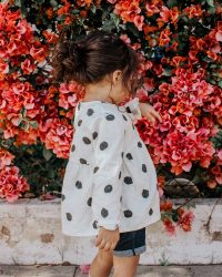 girl in black and white polka-dot long-sleeved blouse and blue shorts standing beside red petaled flowers during daytime